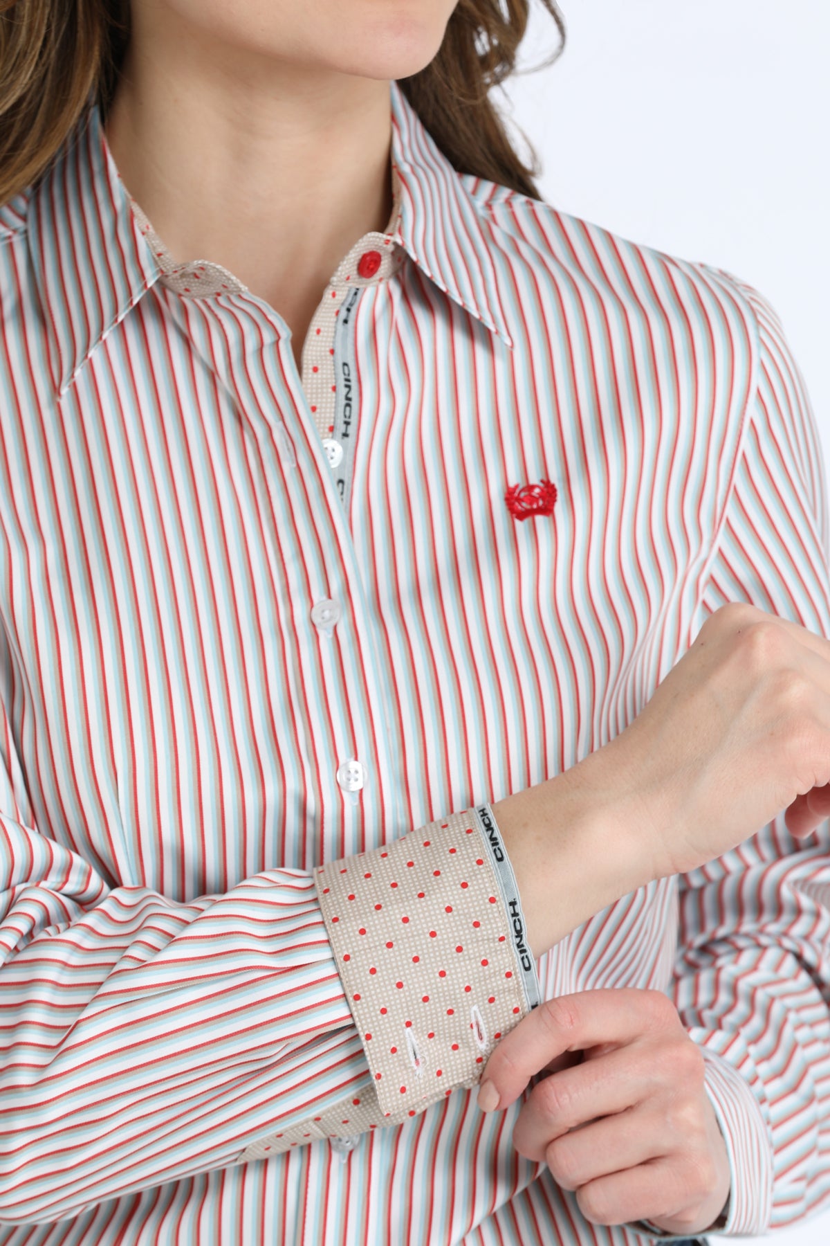 A close-up of a woman wearing a long-sleeve button-down Cinch shirt with thin red and white vertical stripes, featuring a small red embroidered heart on the chest and a patterned cuff.