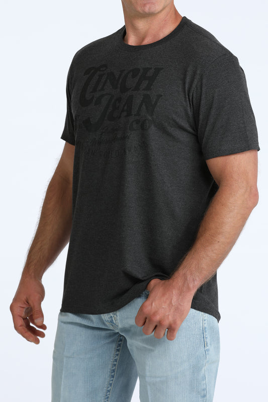 A man wearing a dark charcoal gray heathered t-shirt with a tonal black graphic print on the front that reads 'Cinch Jean Co', paired with light blue jeans, against a plain white background.