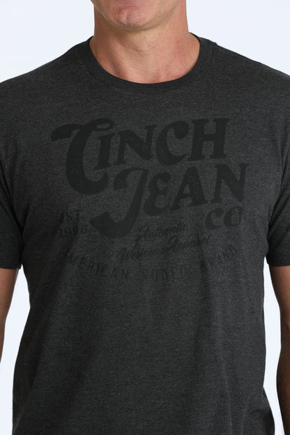 A close-up of a man wearing a dark heather gray t-shirt with a faded black graphic print that reads 'Cinch Jean Co'.