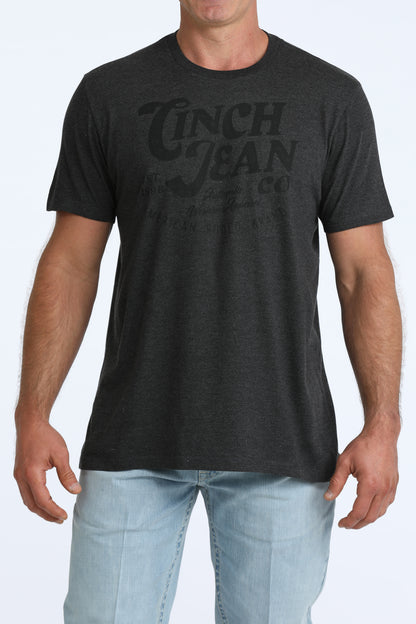 A man wearing a dark charcoal gray heather t-shirt with a faded black graphic print that reads 'CINCH JEAN co.' on the chest, paired with light blue jeans against a light gray background.