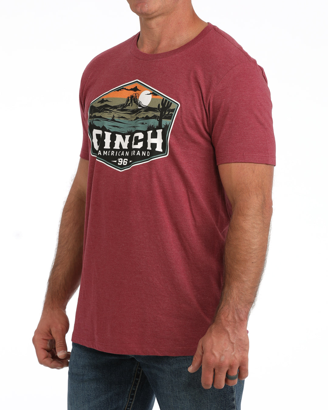 A man wearing a heathered red crewneck t-shirt with a large vintage-style graphic on the chest that reads 'CINCH' with a landscape illustration, paired with blue jeans against a white background.