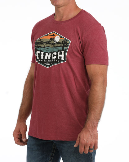A man wearing a heathered red crewneck t-shirt with a large vintage-style graphic on the chest that reads 'CINCH' with a landscape illustration, paired with blue jeans against a white background.
