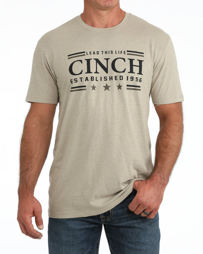 Man wearing a kahki Cinch tshirt and blue jeans on a white background