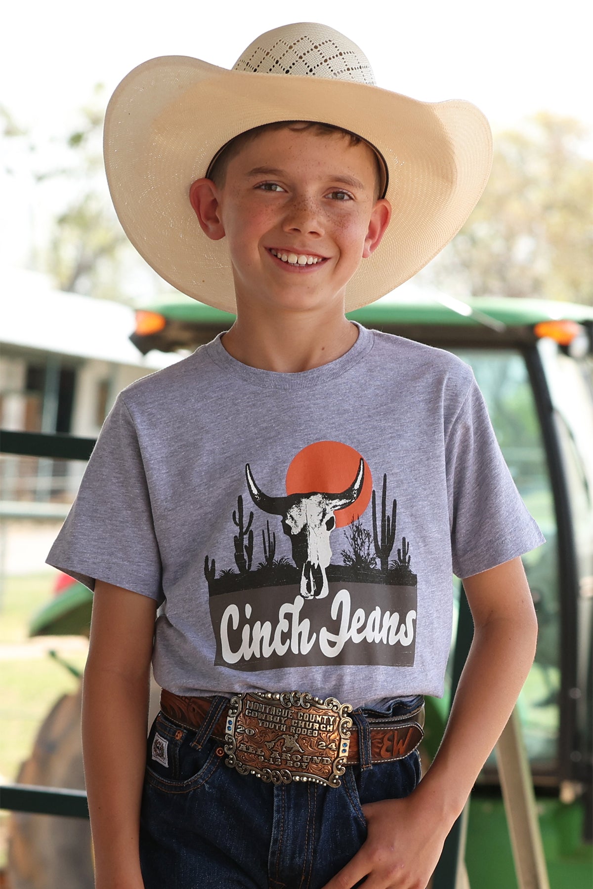 Young boy wearing a Cinch Jeans t-shirt with a cowboy hat and belt, standing in front of a blurred background.