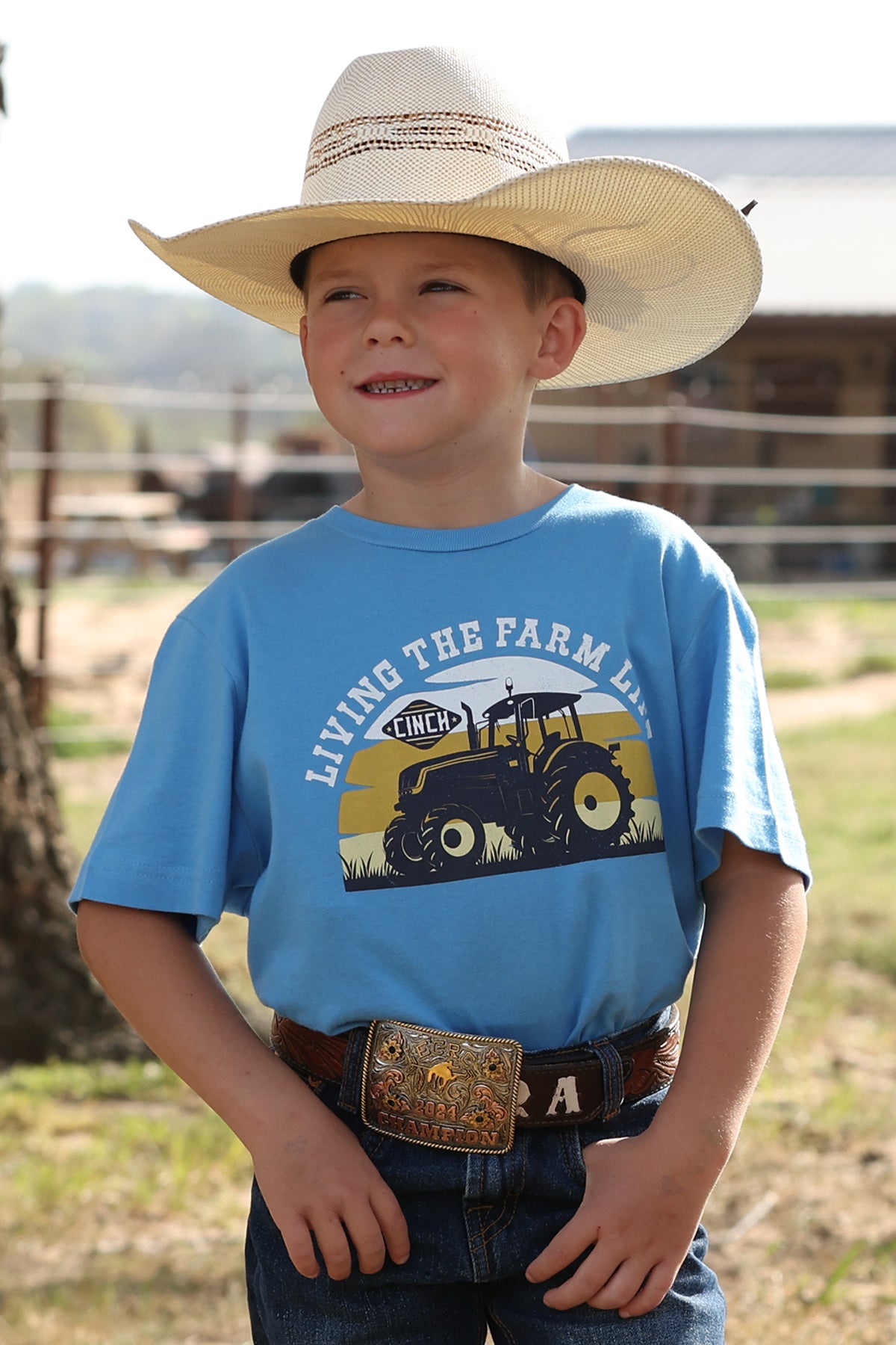 Child wearing a cowboy hat and blue cinch t-shirt with a tractor graphic, standing outdoors.
