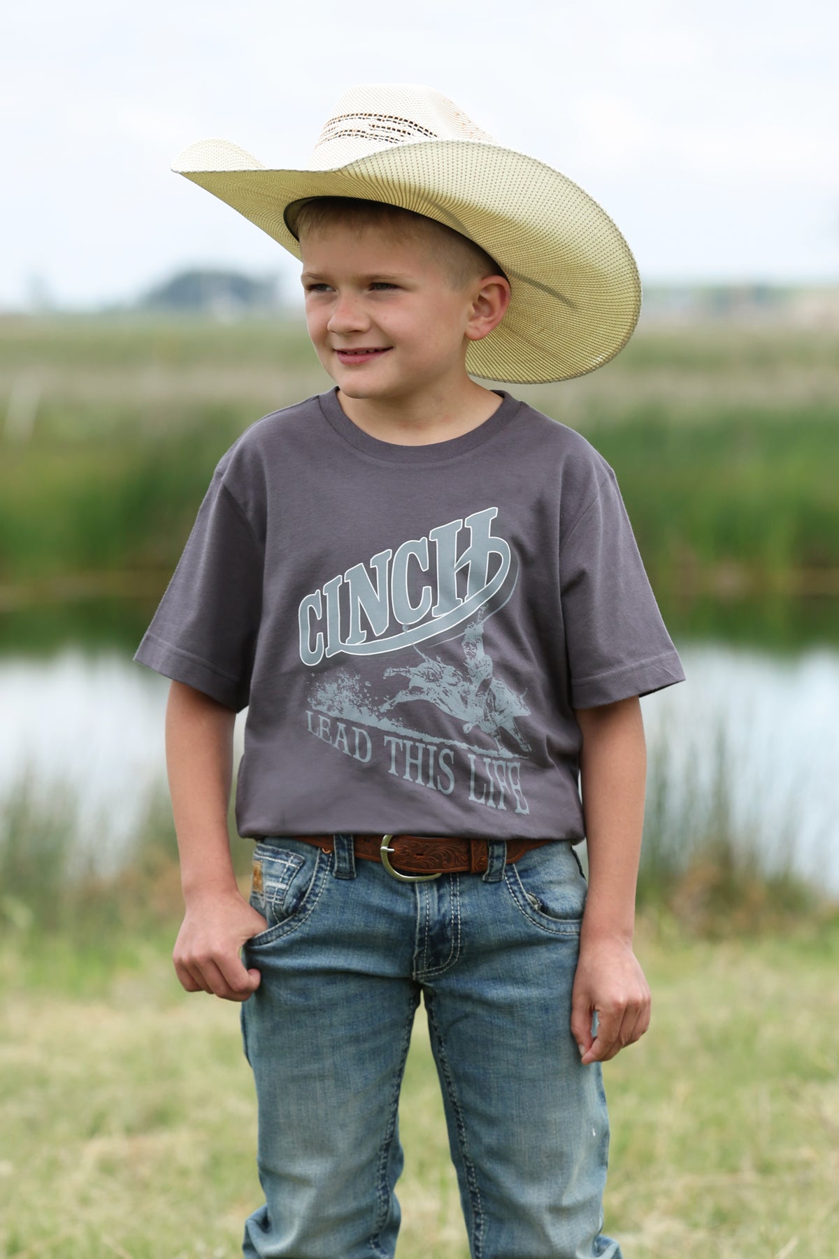 Young boy wearing a cowboy hat and gray t-shirt with 'Cinch' branding outdoors.