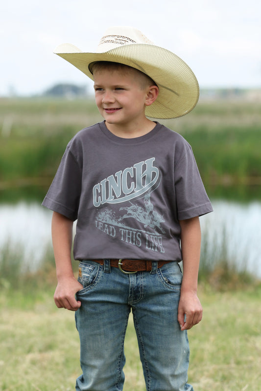 Young boy wearing a cowboy hat and gray t-shirt with 'Cinch' branding outdoors.
