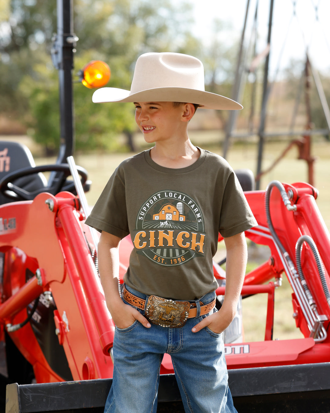 A young boy wearing a beige cowboy hat, an olive green graphic t-shirt with 'CINCH' printed on the front, blue jeans, and a western belt, standing in front of a red tractor.