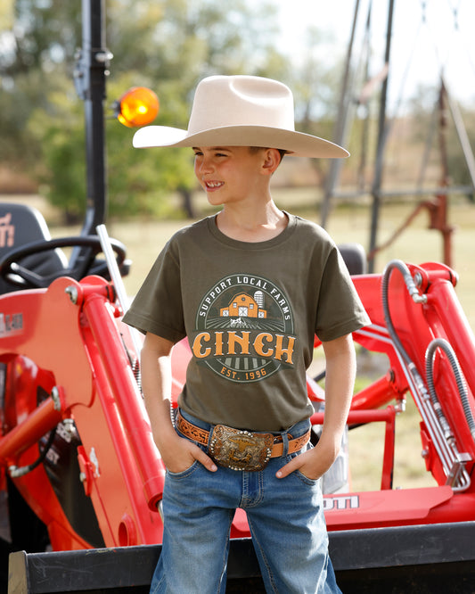 A young boy wearing a beige cowboy hat, an olive green graphic t-shirt with 'CINCH' printed on the front, blue jeans, and a western belt, standing in front of a red tractor.