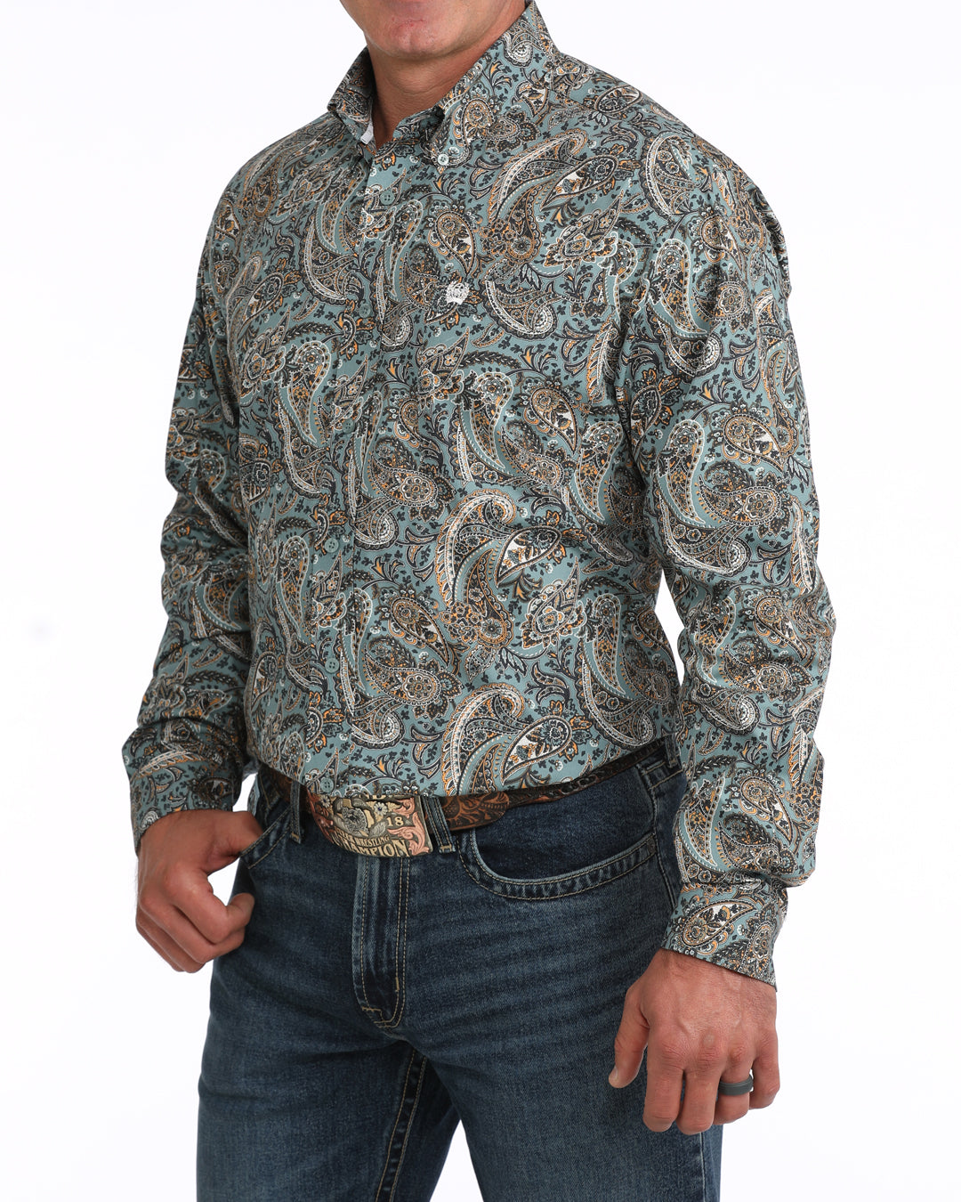 A man wearing a long-sleeve western-style button-up Cinch shirt with an intricate paisley pattern in muted teal, beige, and brown tones, paired with dark blue jeans and a brown belt.
