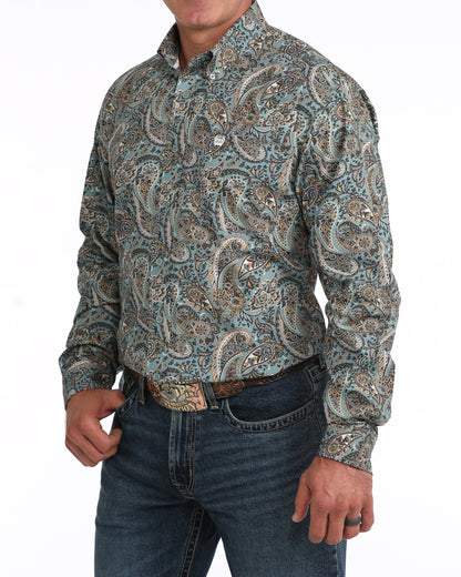 A man wearing a long-sleeve western-style button-up Cinch shirt with an intricate paisley pattern in muted teal, beige, and brown tones, paired with dark blue jeans and a brown belt.