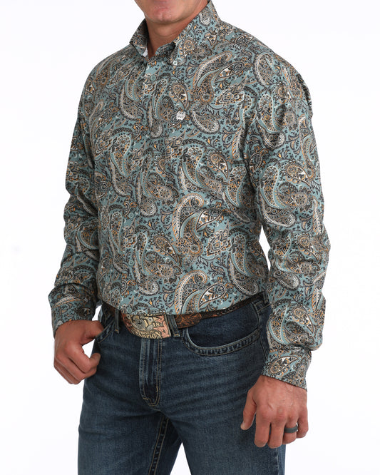 A man wearing a long-sleeve western-style button-up Cinch shirt with an intricate paisley pattern in muted teal, beige, and brown tones, paired with dark blue jeans and a brown belt.