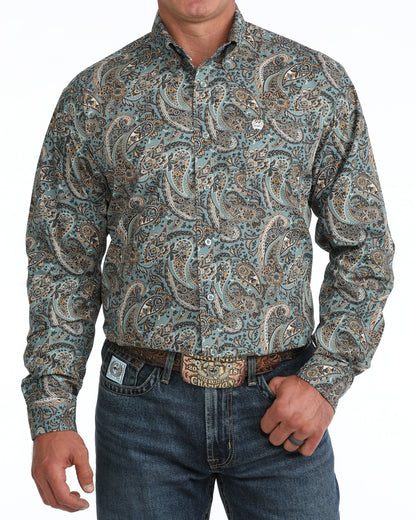 A man wearing a long-sleeve western Cinch shirt with a detailed paisley pattern in teal, brown, and beige, paired with blue jeans and a western belt.