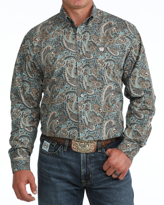 A man wearing a long-sleeve western Cinch shirt with a detailed paisley pattern in teal, brown, and beige, paired with blue jeans and a western belt.
