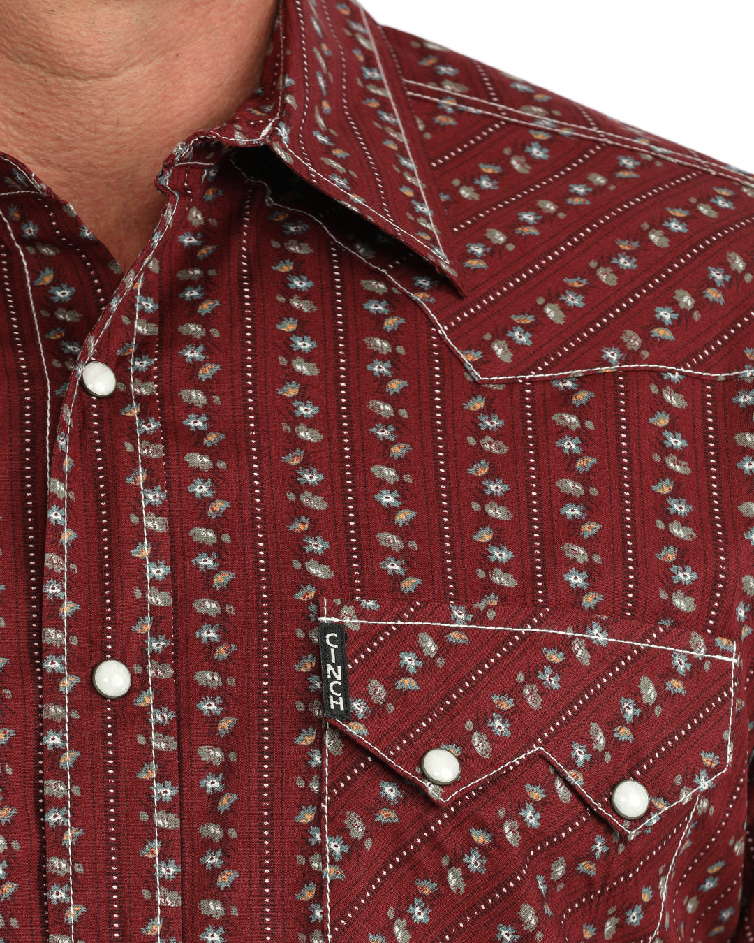 Close-up of a men's western-style button-up Cinch shirt with a deep collar, chest flap pocket, and white snap buttons. The fabric is a dark red/burgundy base with a repeating vertical pattern of small white and gray floral or leaf motifs.