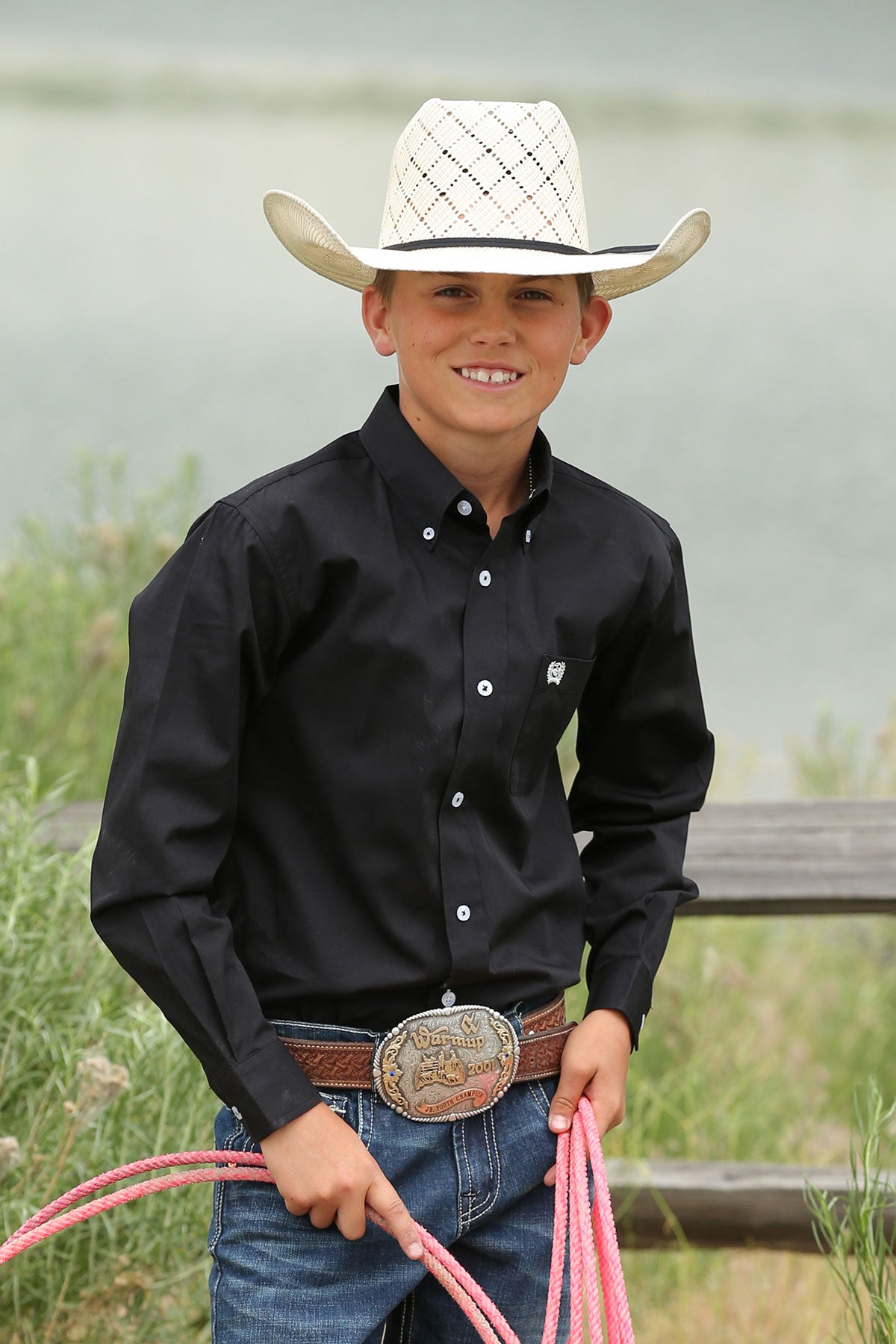 boy wearing a black Cinch shirt, blue jeans and a belt and a white cowboy hat holding a pink rope standing next to a fence