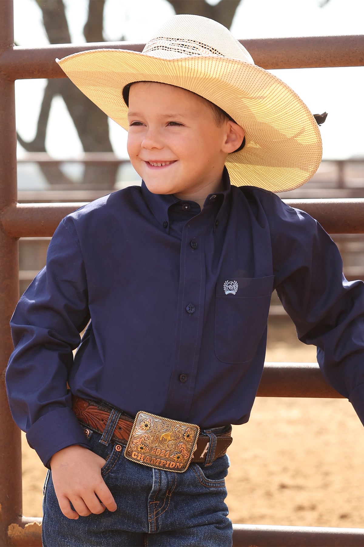 Child wearing a navy blue cinch shirt and cowboy hat with a belt, standing in front of a wooden fence.