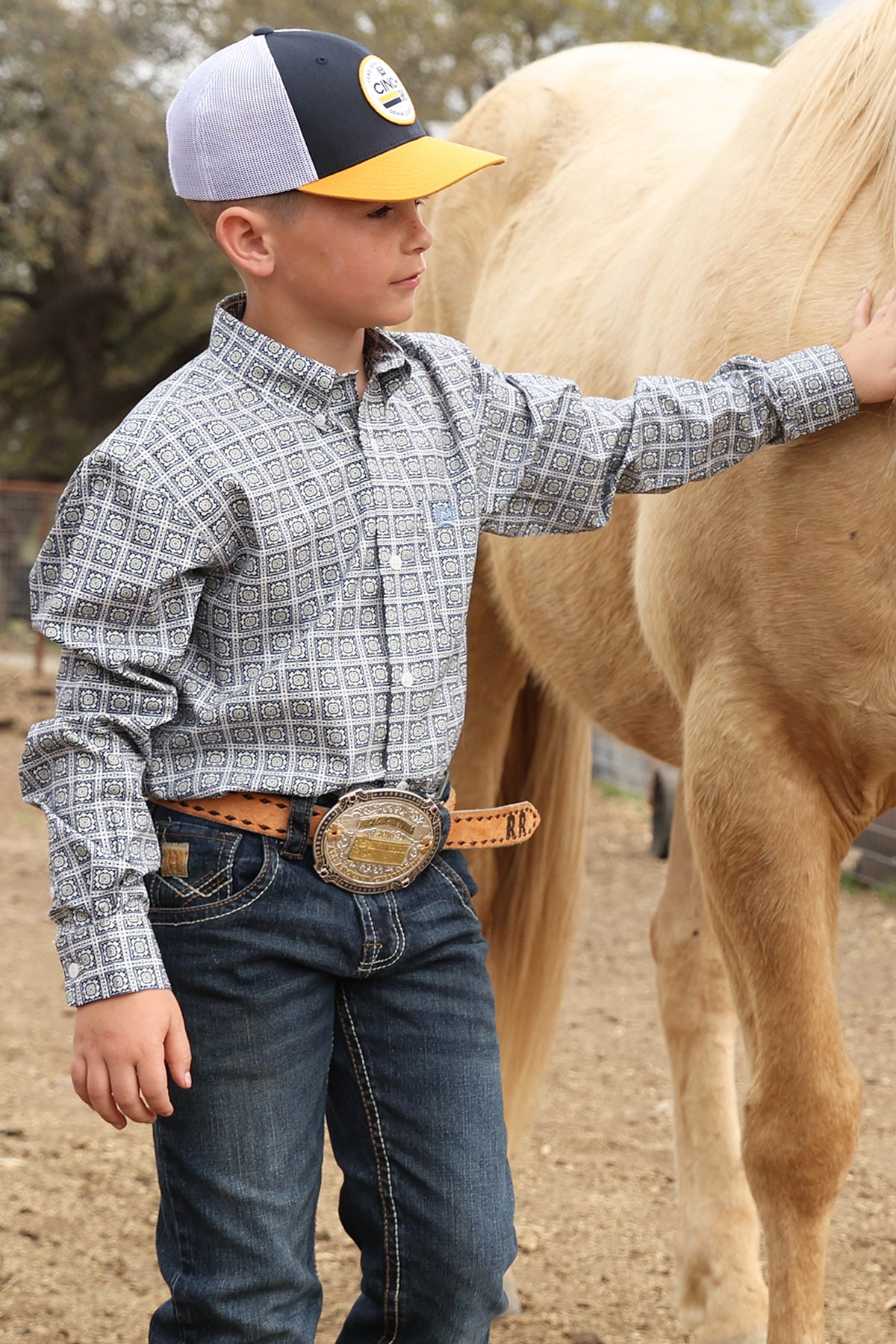Boy wearing a white and navy Cinch shirt, blue jeans and  ball cap petting a horse