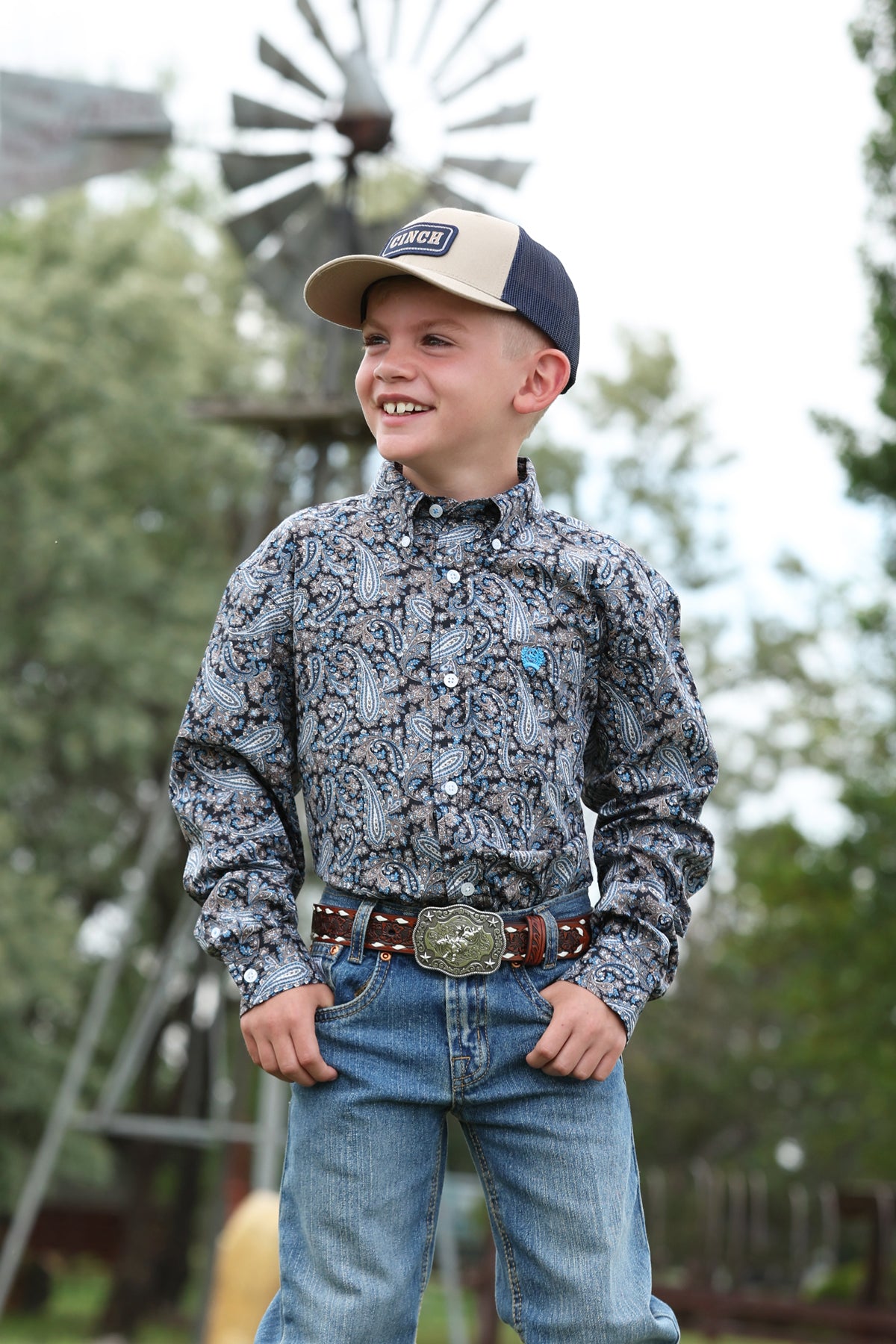 Young boy in a patterned Cinch shirt and jeans with a cap, standing outdoors with a windmill in the background.
