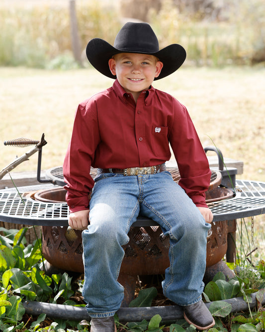 Young boy in cowboy attire sitting on a rustic metal object outdoors. Wearing a red Cinch shirt