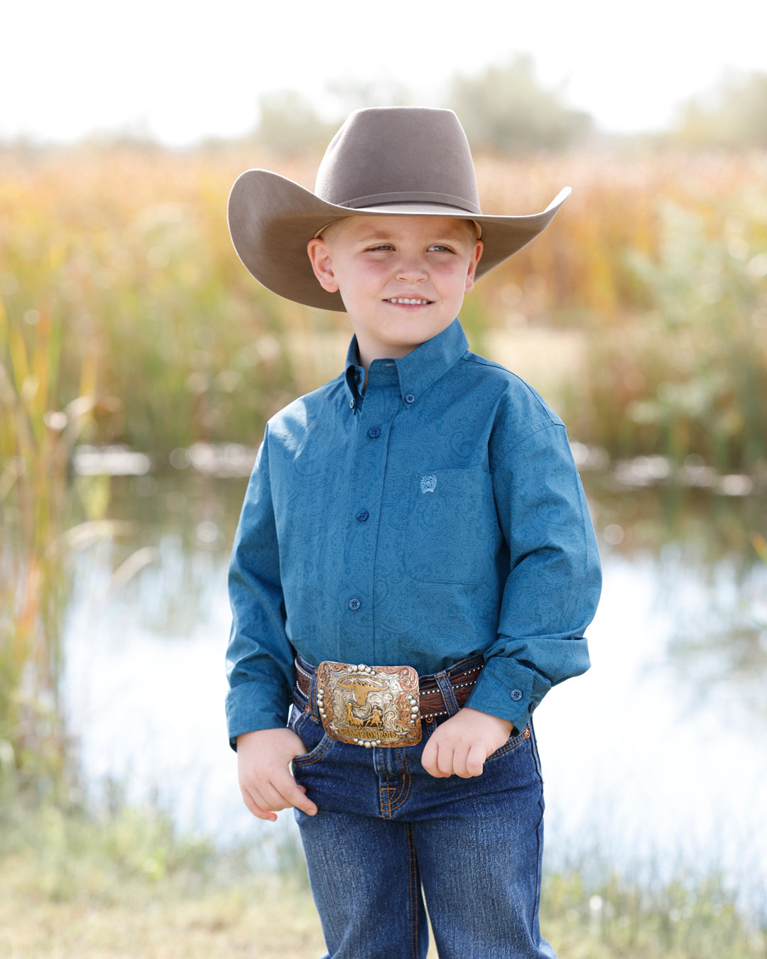 Little boy wearing a teal paisley Cinch shirt, blue jeans, a buckle and a grey cowboy hat with a pond and grass behind him