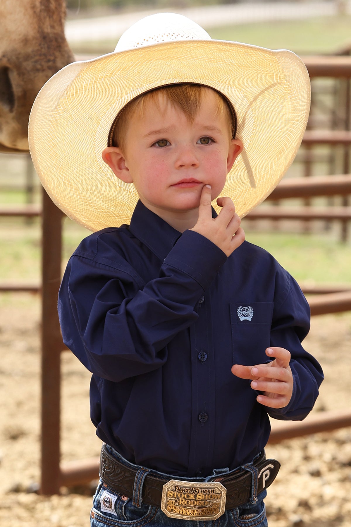 Young boy wearing a straw cowboy hat and navy cinch shirt with a logo, standing in an outdoor setting.