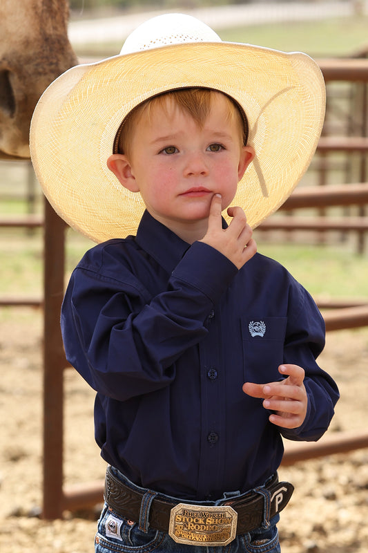 Young boy wearing a straw cowboy hat and navy cinch shirt with a logo, standing in an outdoor setting.