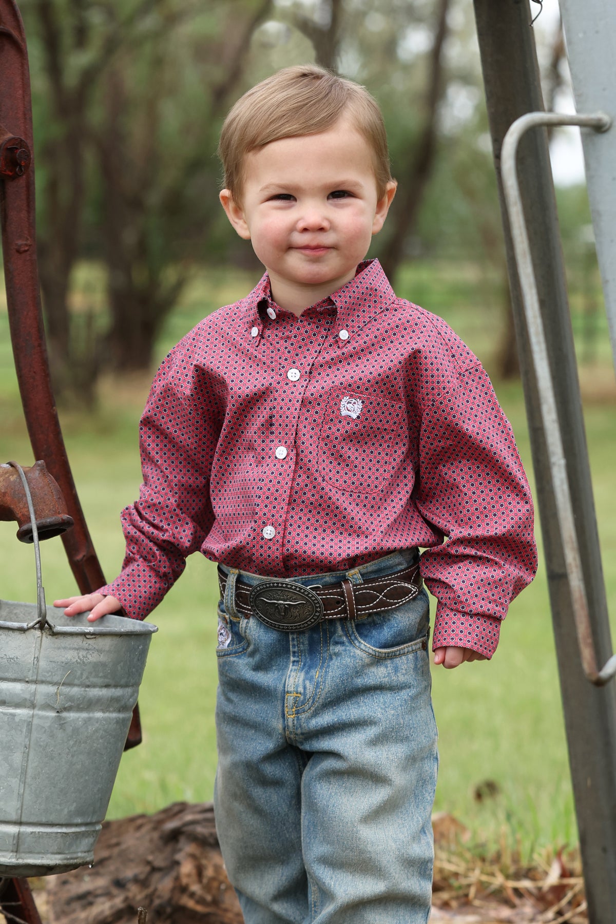 Child wearing a red patterned Cinch  shirt and blue jeans standing outdoors near a metal bucket and ladder.