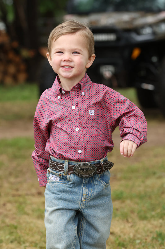 Child wearing a red patterned Cinch shirt and blue jeans standing outdoors with a vehicle in the background.