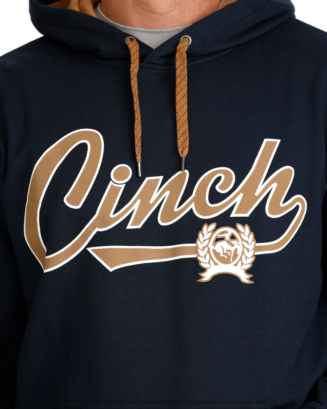 Close-up of a navy pullover hoodie with a large gold and white 'Cinch' script graphic across the chest, plus a matching gold crest emblem. The hoodie has tan drawstrings and a gray undershirt visible at the collar.