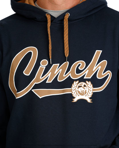 Close-up of a navy pullover hoodie with a large gold and white 'Cinch' script graphic across the chest, plus a matching gold crest emblem. The hoodie has tan drawstrings and a gray undershirt visible at the collar.