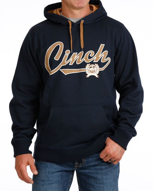 A man wearing a navy blue pullover hoodie with a large gold 'Cinch' script logo across the chest, paired with blue jeans against a plain white background.