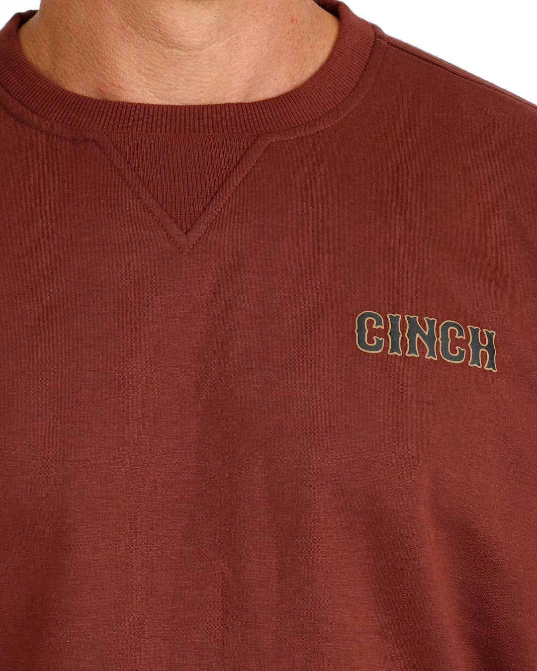 Close-up of a brown crewneck sweatshirt with the brand name 'CINCH' embroidered in green and white on the left chest, worn by a model.