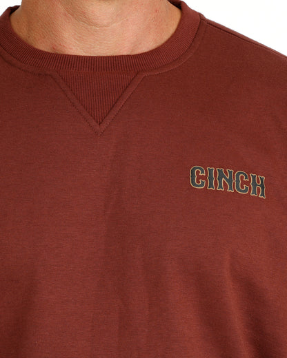 Close-up of a brown crewneck sweatshirt with the brand name 'CINCH' embroidered in green and white on the left chest, worn by a model.