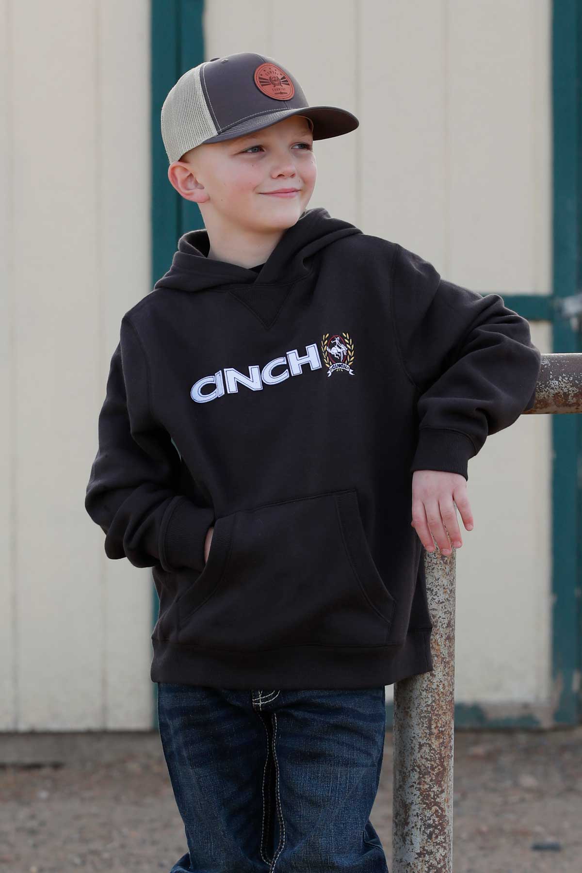 Boy wearing a brown Cinch sweater, blue jeans and ball cap leaning on a post