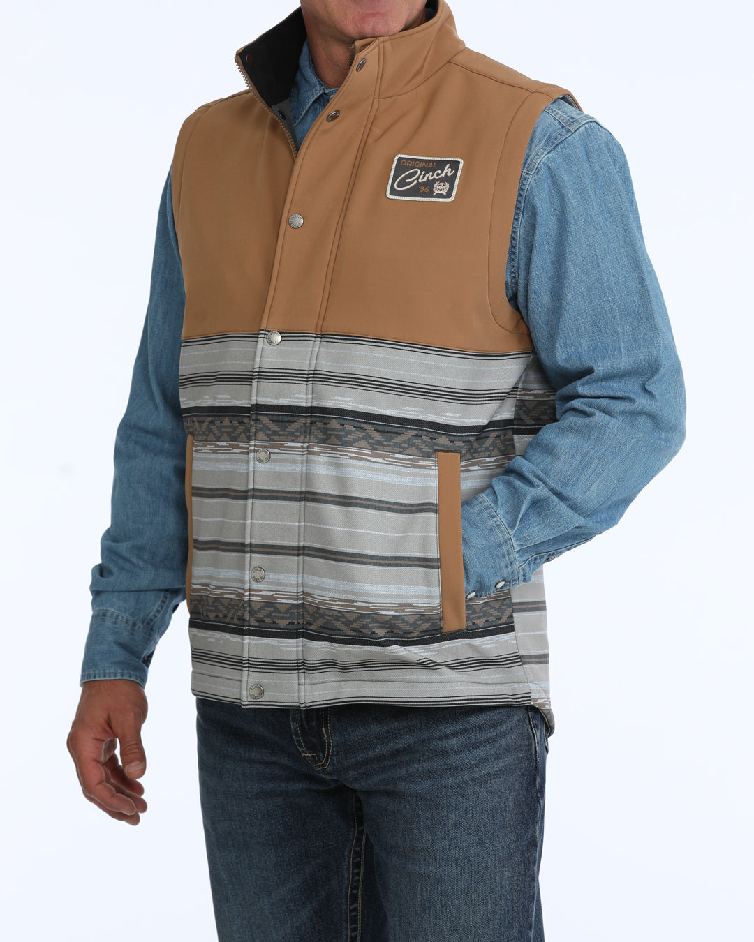 Man wearing a tan and grey Cinch vest with a denim shirt and blue jeans on a white background