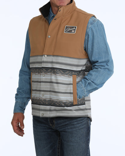 Man wearing a tan and grey Cinch vest with a denim shirt and blue jeans on a white background