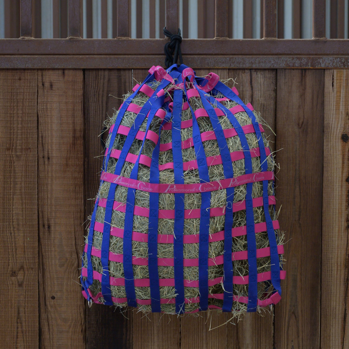 Professional's choice Hay bag with pink and blue straps hanging on a wooden fence