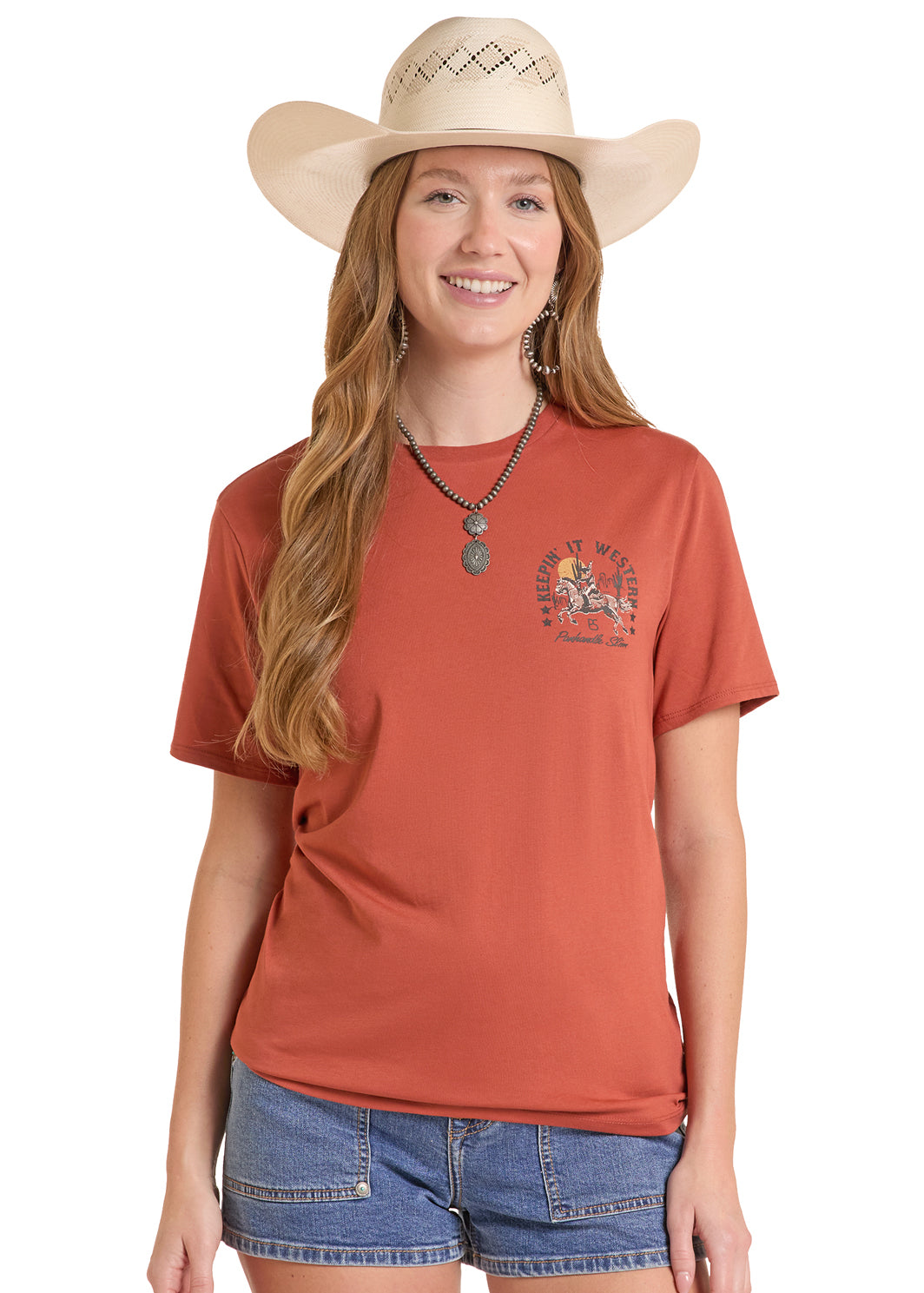 Woman wearing a rust-colored Panhandle Slim t-shirt with a graphic design, denim shorts, and a beige cowboy hat on a white background.