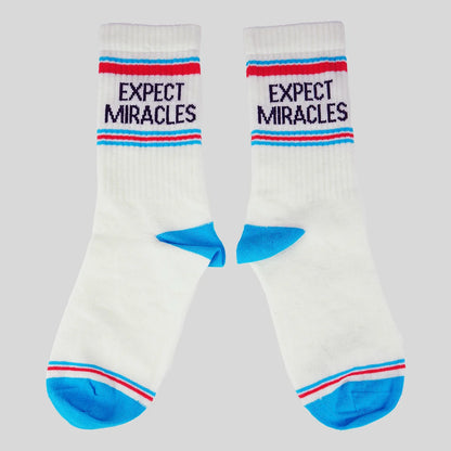A pair of white crew socks from Kerusso with blue and red stripes, featuring the text 'EXPECT MIRACLES' in navy blue on the upper cuff, and bright blue heels and toes, set against a solid gray background.