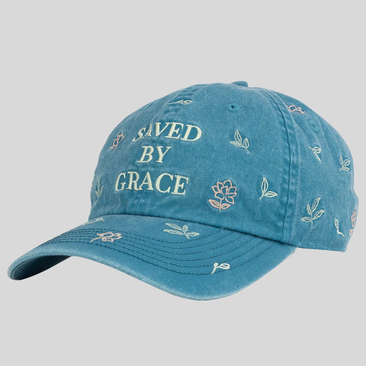 A blue denim baseball cap from Kerusso with 'SAVED BY GRACE' embroidered on the front and small floral and leaf motifs scattered across the fabric, shown against a solid gray background.