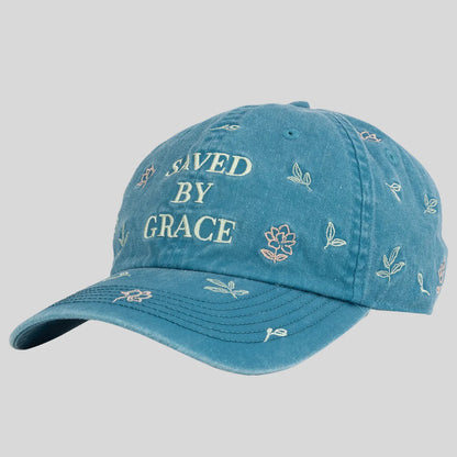 A blue denim baseball cap from Kerusso with 'SAVED BY GRACE' embroidered on the front and small floral and leaf motifs scattered across the fabric, shown against a solid gray background.