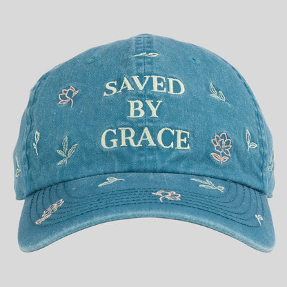 A blue distressed baseball cap from Kerusso with 'SAVED BY GRACE' embroidered in cream on the front, surrounded by small floral and leaf motifs, shown against a light gray background.