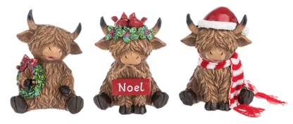 Three Highland cow Christmas figurines from Ganz on a white background. The left cow holds a green wreath with a red bow.