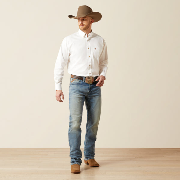 Man wearing a white Ariat shirt, blue jeans, and a brown cowboy hat on a plain background