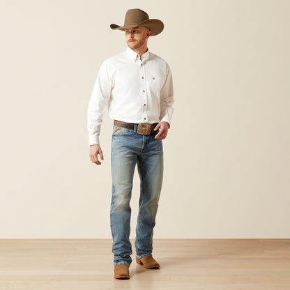 Man wearing a white Ariat shirt, blue jeans, and a brown cowboy hat on a plain background