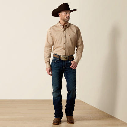 A man wearing a tan Ariat long-sleeve button-down shirt, dark blue jeans, a brown cowboy hat, and brown boots, standing in a studio setting with a beige wall and light wood floor.