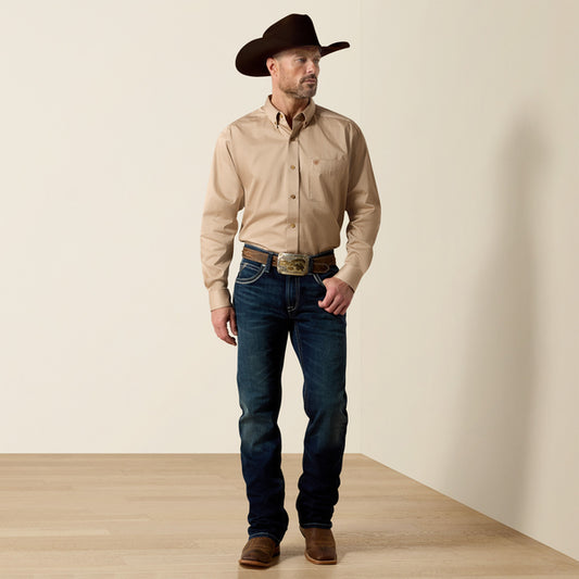 A man wearing a tan Ariat long-sleeve button-down shirt, dark blue jeans, a brown cowboy hat, and brown boots, standing in a studio setting with a beige wall and light wood floor.