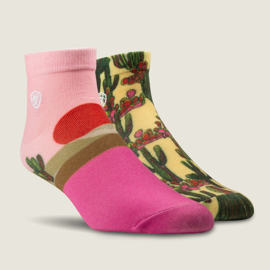 A pair of Ariat ankle socks displayed on a light beige background. The front sock is bright pink with a red and brown circular graphic and green cactus print near the cuff.