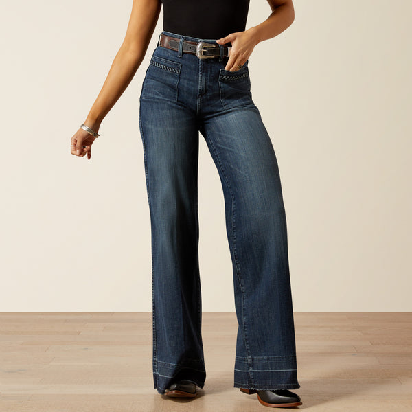 woman wearing blue Ariat jeans, a black t shirt and cowboy boots on a cream background